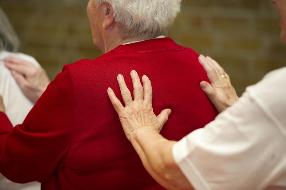 Nahaufnahme von zwei Händen, die sanft die Schultern einer älteren Frau in rotem Pullover berühren.