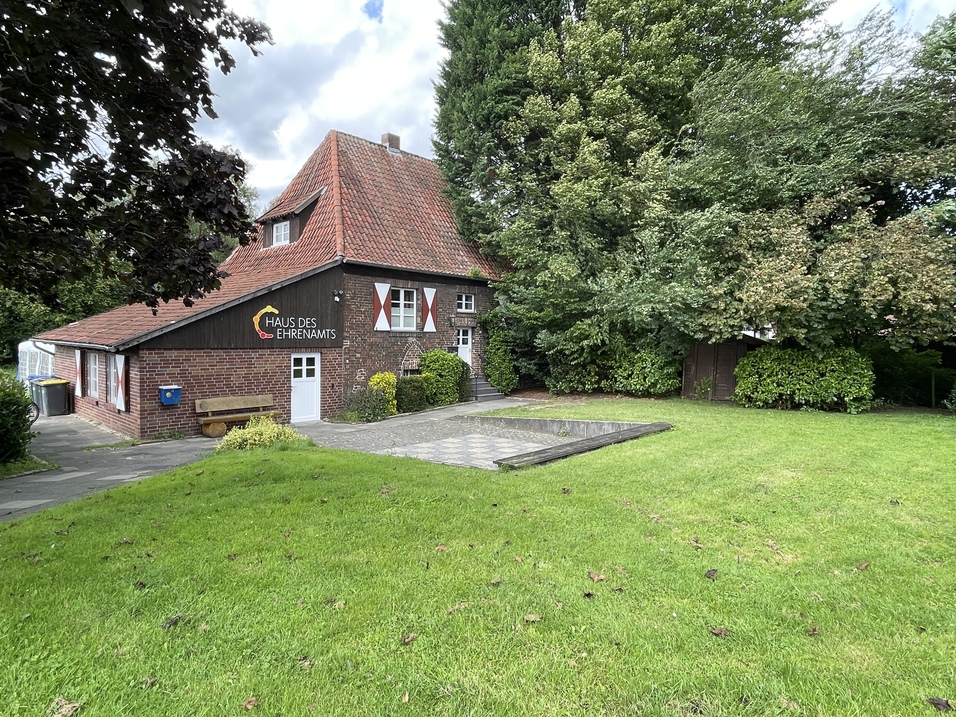 Haus des Ehrenamts mit rotem Satteldach in einem gr&uuml;nen Garten, umgeben von B&auml;umen und einer gepflasterten Fl&auml;che.