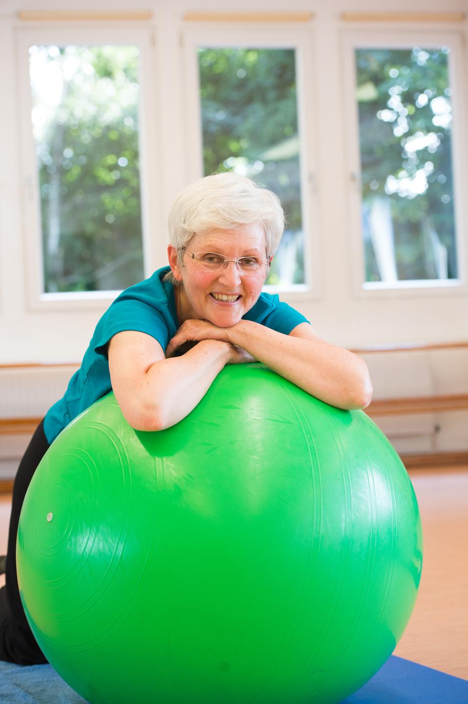 Lächelnde ältere Frau lehnt auf einer großen grünen Gymnastikball in einem hellen Raum mit Fenstern.
