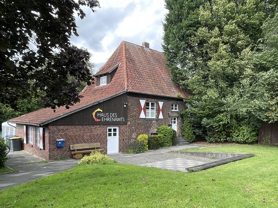 Haus des Ehrenamts mit rotem Satteldach in einem grünen Garten, umgeben von Bäumen und einer gepflasterten Fläche.