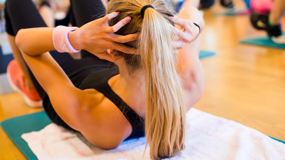 Frau macht Crunches auf einer Matte im Fitnessstudio, während andere Teilnehmer im Hintergrund trainieren.