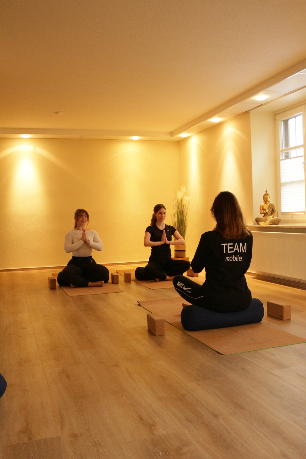 Drei Frauen in Yogastellungen sitzen auf Matten, umgeben von Yoga-Bl&ouml;cken in einem hellen Raum.