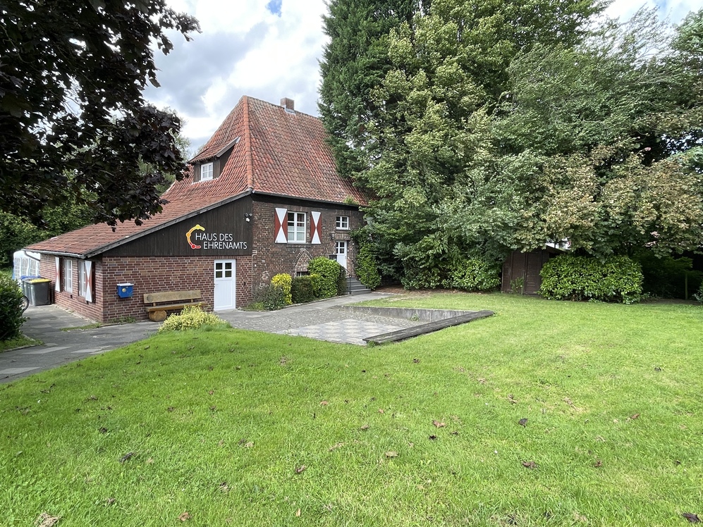 Haus des Ehrenamts mit rotem Satteldach in einem gr&uuml;nen Garten, umgeben von B&auml;umen und einer gepflasterten Fl&auml;che.