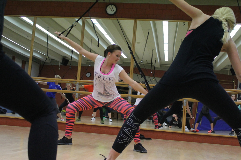 Sicht auf eine Tanz-Workout-Gruppe mit einer Frau in pinken Streifenleggings und T-Shirt vor einem Spiegel.