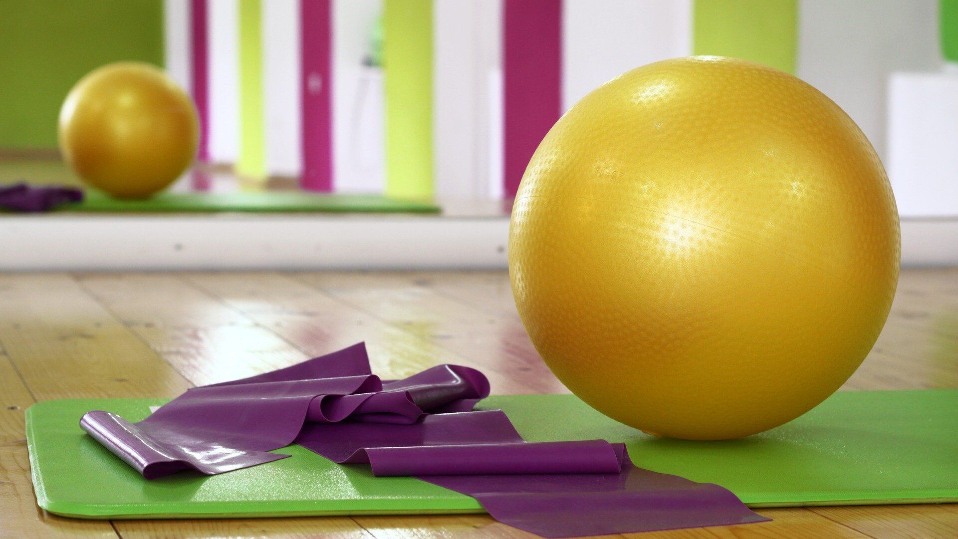 Gelbe Übungsball und purple Widerstandsbänder liegen auf einer grünen Yogamatte in einem hellen Raum mit bunten Wänden.