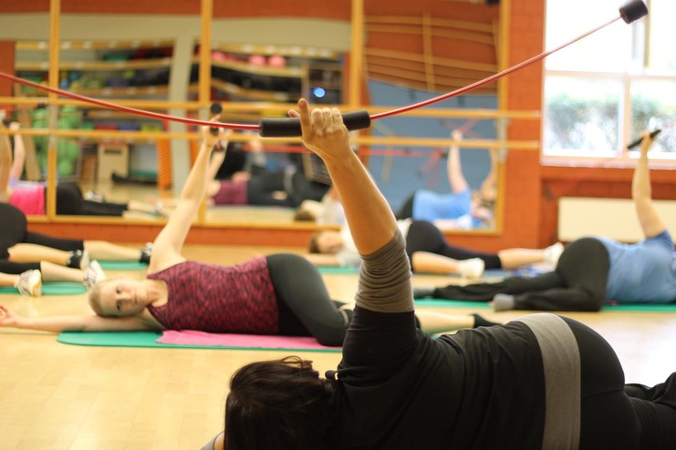 Frauen in Sportkleidung liegen auf Matten und nutzen Widerstandsbänder in einem Fitnessraum mit Spiegeln.
