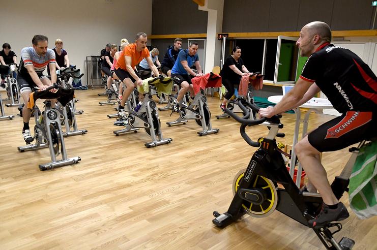 Gruppentraining mit mehreren Personen auf Spinning-Bikes in einem Fitnessraum, ein Trainer gibt Anweisungen.