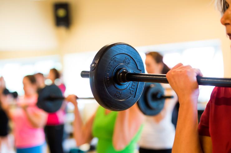 Nahaufnahme einer Hantelstange, die von einer Person in Sportbekleidung im Fitnessstudio angehoben wird.