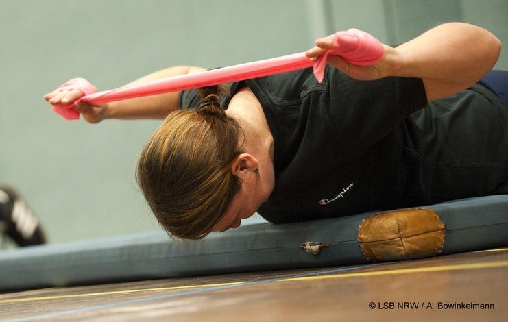 Frau dehnt sich mit einem roten Widerstandsband auf einer Trainingsmatte, Kopf gesenkt, in sportlicher Kleidung.