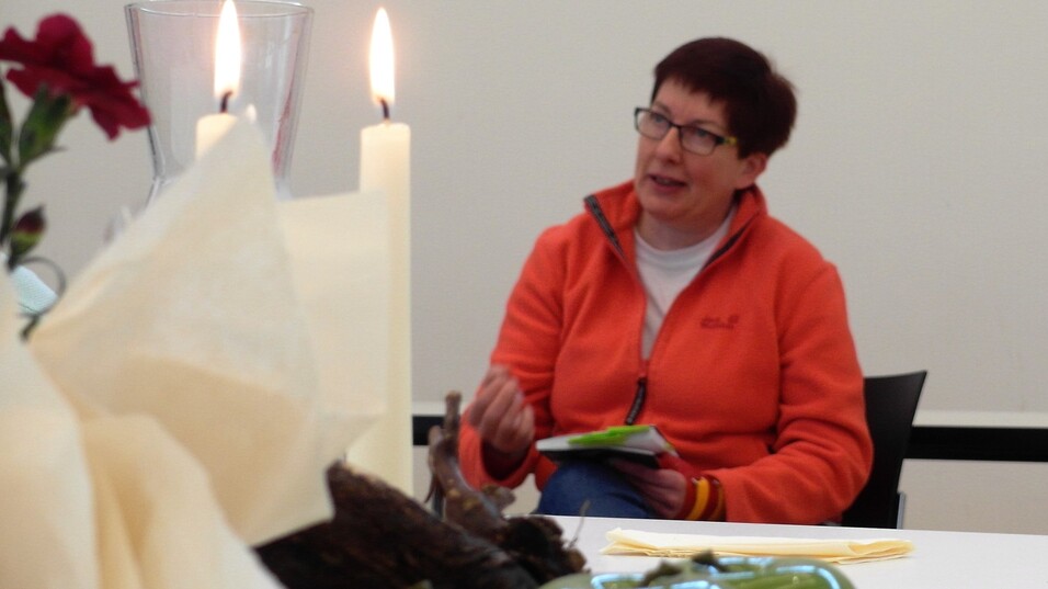 Eine Frau mit Brille in orangefarbener Jacke sitzt an einem Tisch, umgeben von Blumen und Kerzen.