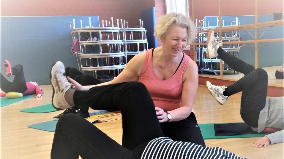 Eine Trainerin unterstützt eine Teilnehmerin beim Beinheben auf einer Ümatte in einem Fitnessraum.