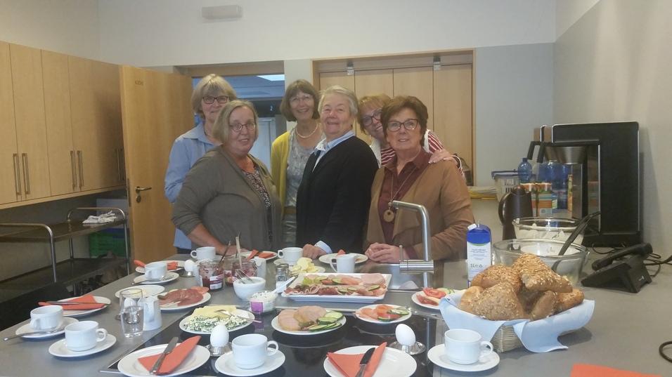 Gruppe von sechs Frauen lächelt vor einem reichhaltig gedeckten Frühstückstisch in einer modernen Küche.