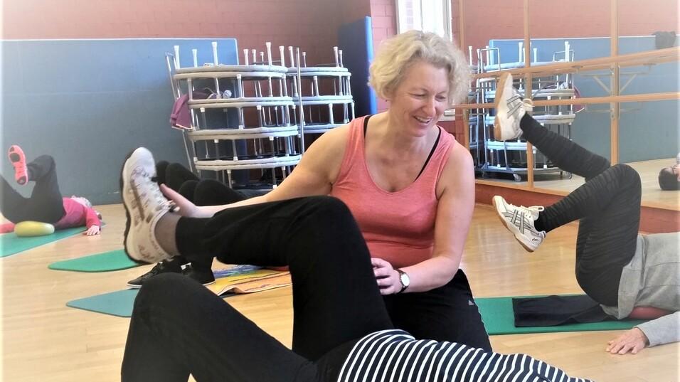 Eine Trainerin unterstützt eine Teilnehmerin beim Beinheben auf einer Ümatte in einem Fitnessraum.