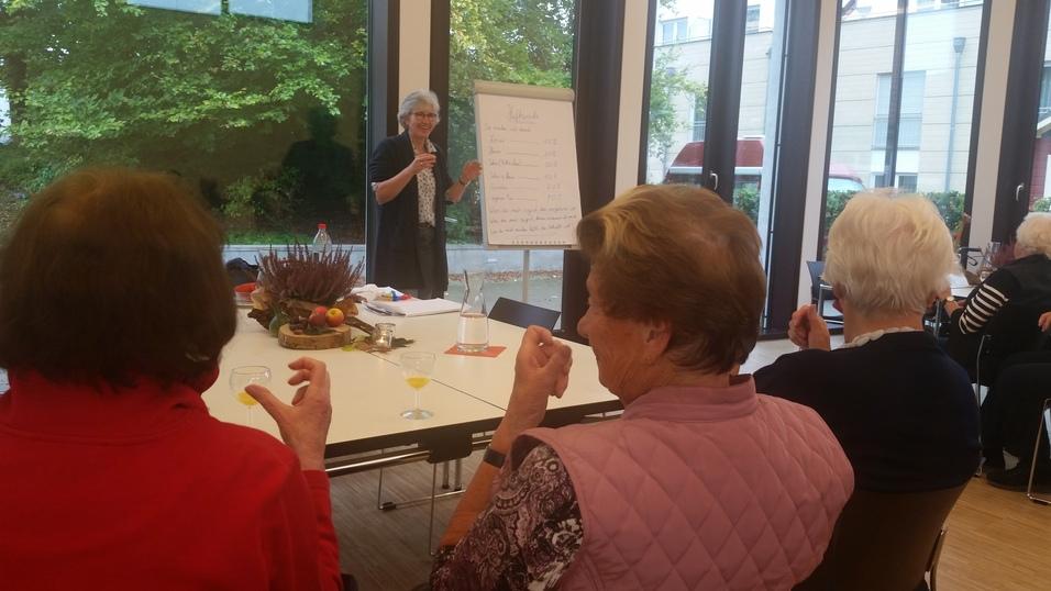 Eine ältere Frau präsentiert vor einem Flipchart, während Teilnehmer mit Getränken am Tisch sitzen.