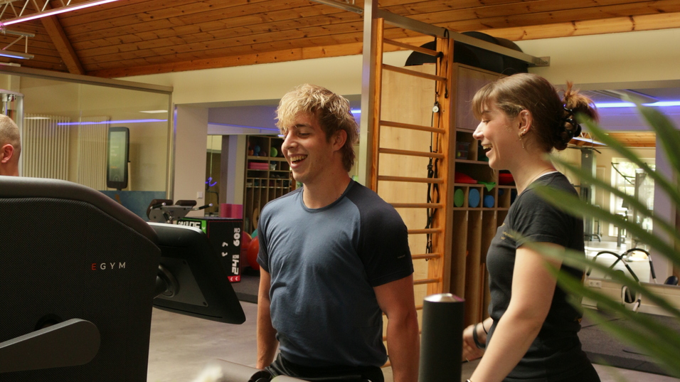 Zwei Personen lächeln und stehen vor einem Fitnessgerät in einem modernen Trainingsraum mit Holzdecke.