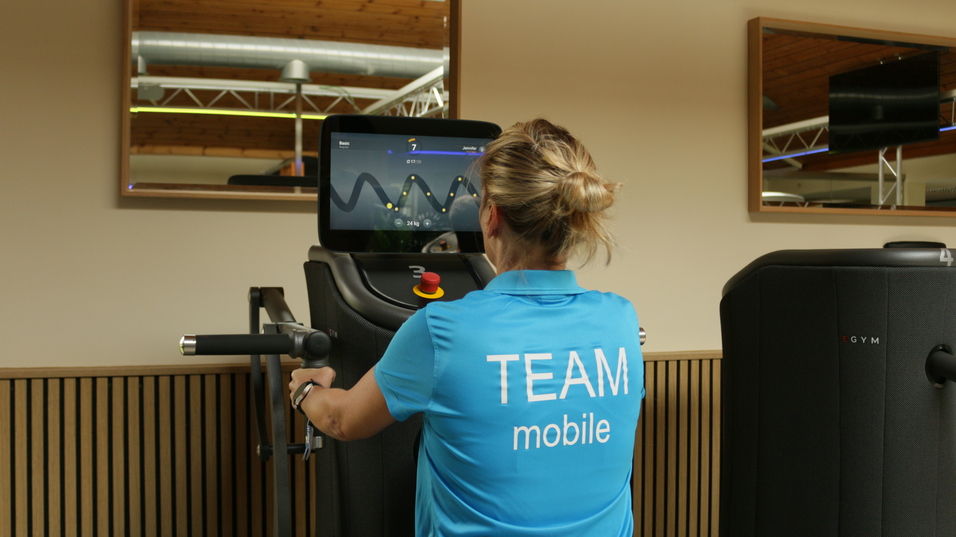 Frau in blauer Sportkleidung trainiert an einem Fitnessgerät mit Bildschirm im Hintergrund.