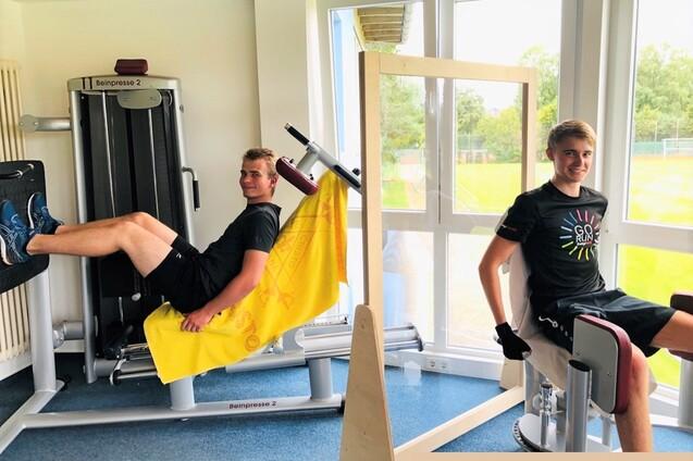 Zwei junge Männer sitzen an Fitnessgeräten in einem hellen Raum mit großen Fenstern und Blick ins Freie.