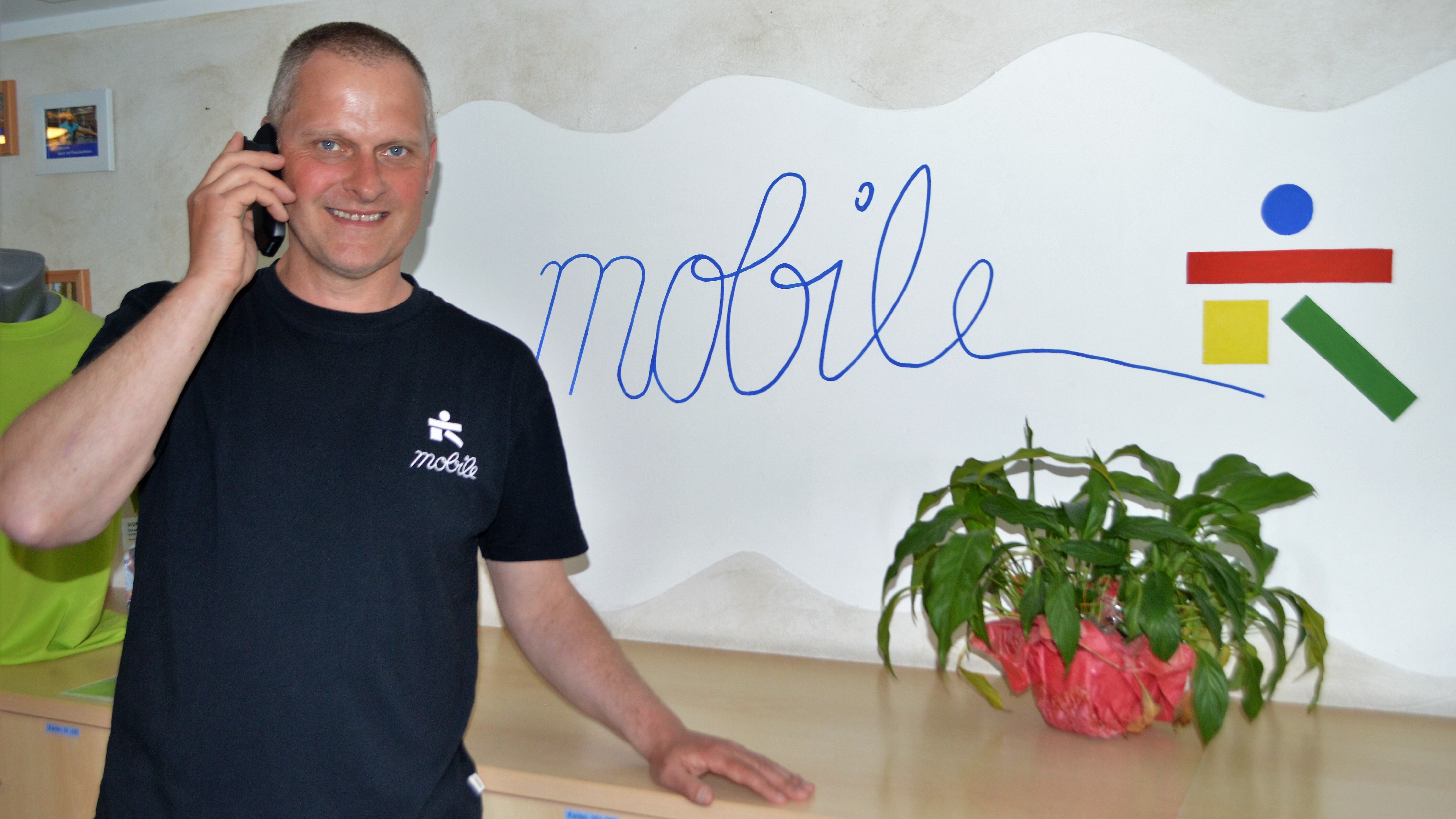 Lächelnder Mann mit Handy vor einer Wand mit dem Schriftzug "mobile" und einer Topfpflanze auf einem Tisch.