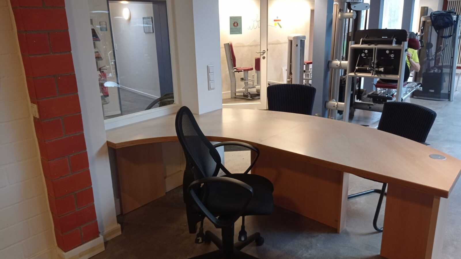 B&uuml;robereich mit kurvenf&ouml;rmigem Schreibtisch und zwei schwarzen St&uuml;hlen, Blick auf angrenzenden Fitnessraum.