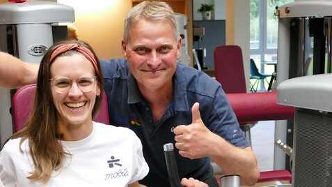 Lächelnde Frau in Sportkleidung sitzt an einem Fitnessgerät, daneben ein Mann mit Daumen hoch. Trainingsumgebung sichtbar.