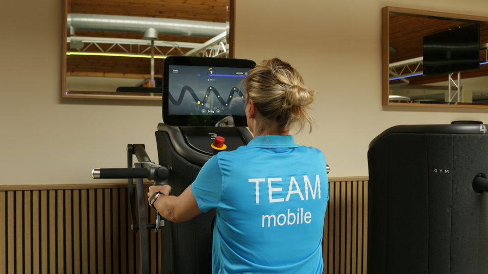 Frau in blauer Sportkleidung trainiert an einem Fitnessgerät mit Bildschirm im Hintergrund.