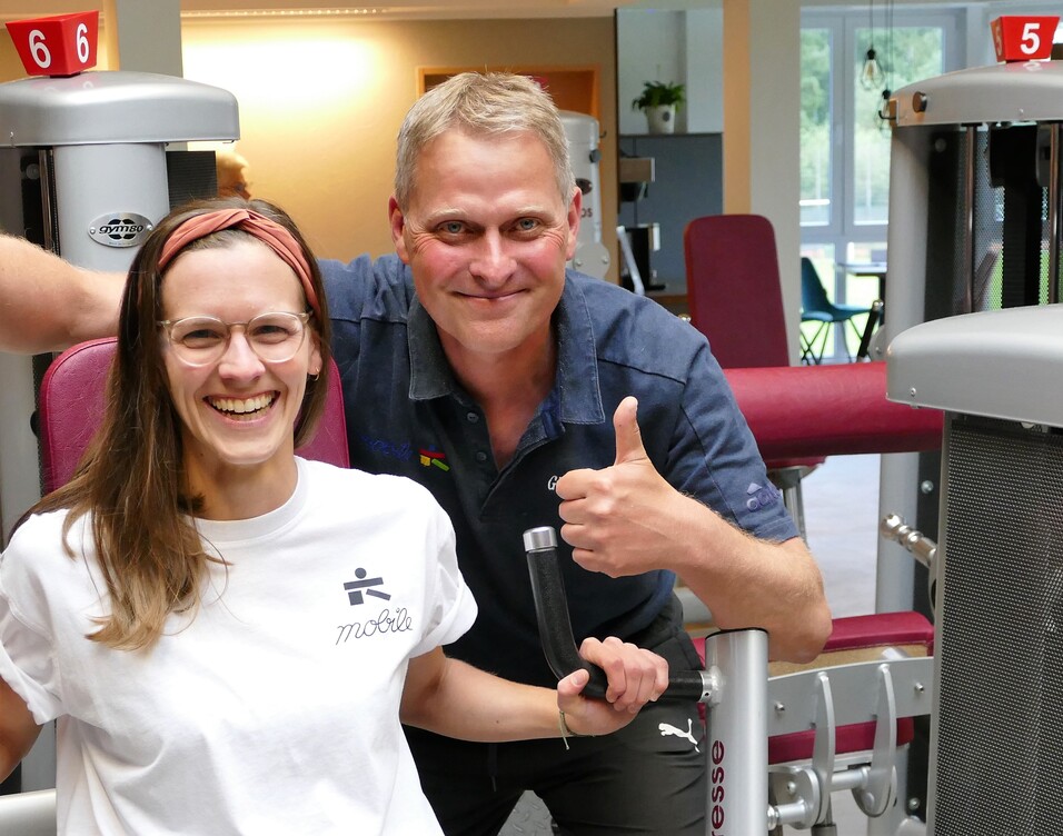 Lächelnde Frau in Sportkleidung sitzt an einem Fitnessgerät, daneben ein Mann mit Daumen hoch. Trainingsumgebung sichtbar.