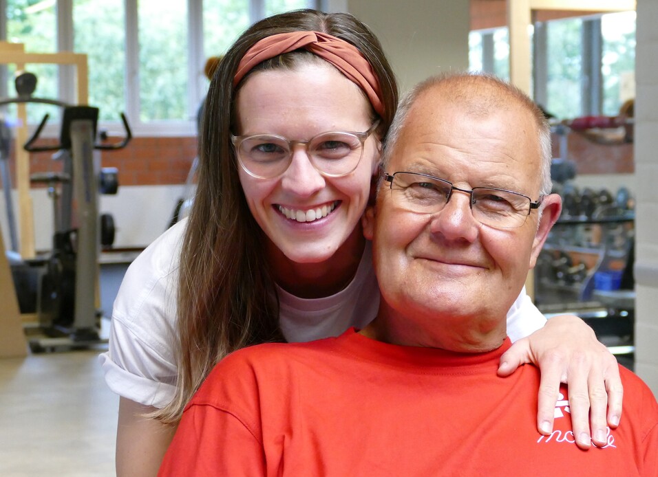 Lächelnde Frau umarmt einen älteren Mann in einem Fitnessstudio. Beide tragen sportliche Kleidung und wirken glücklich.