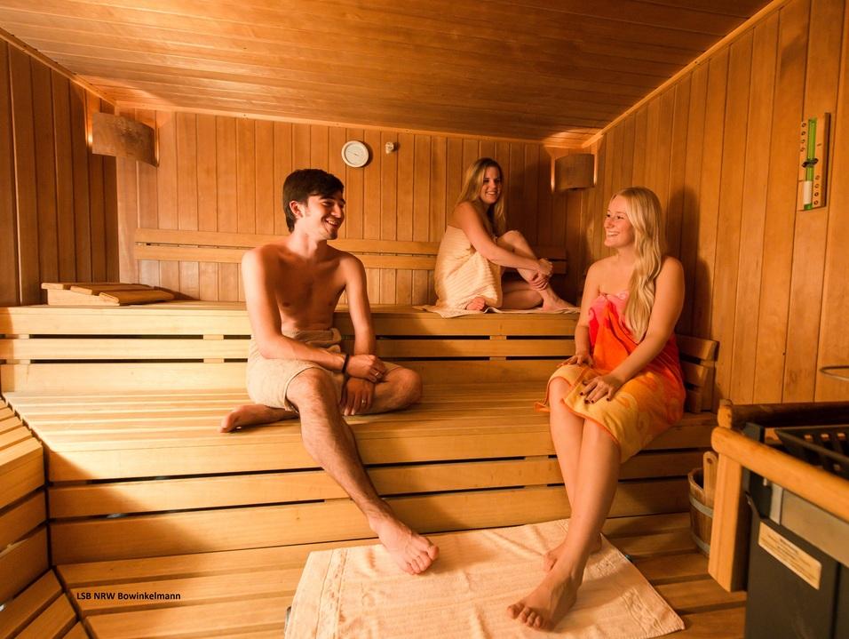Drei Personen in einer Holzsauna sitzen entspannt auf den Bänken, lächelnd und in Handtücher gehüllt.