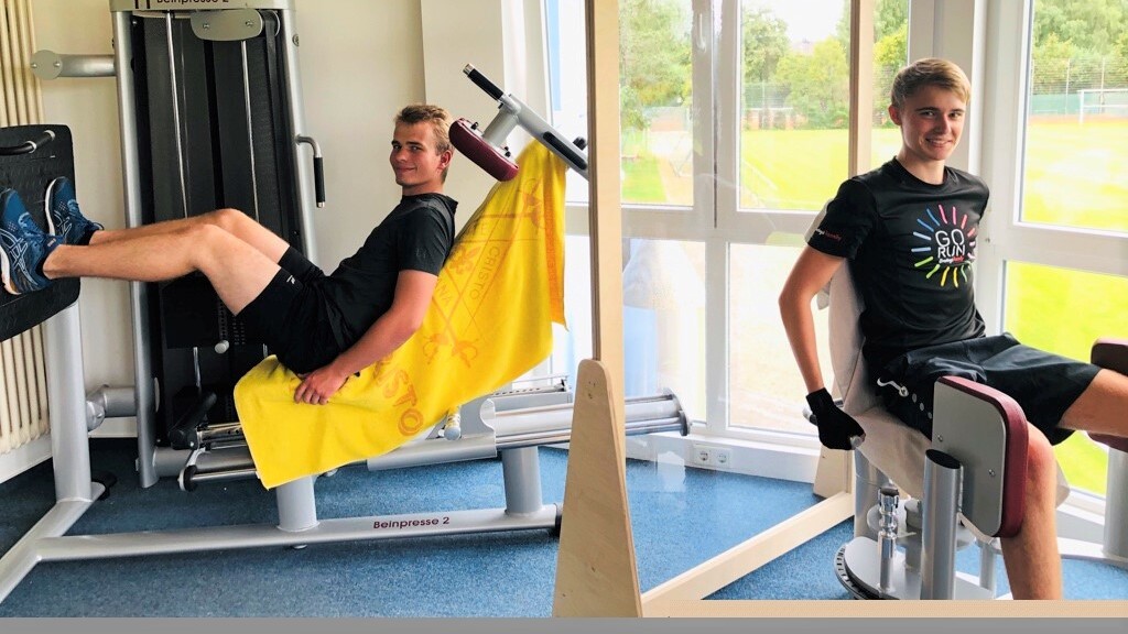 Zwei junge Männer sitzen an Fitnessgeräten in einem hellen Raum mit großen Fenstern und Blick ins Freie.