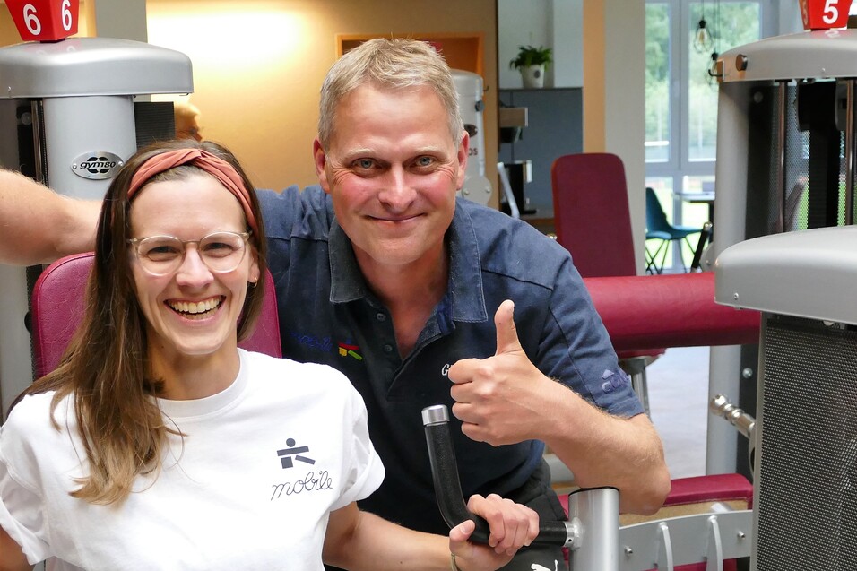 L&auml;chelnde Frau in Sportkleidung sitzt an einem Fitnessger&auml;t, daneben ein Mann mit Daumen hoch. Trainingsumgebung sichtbar.