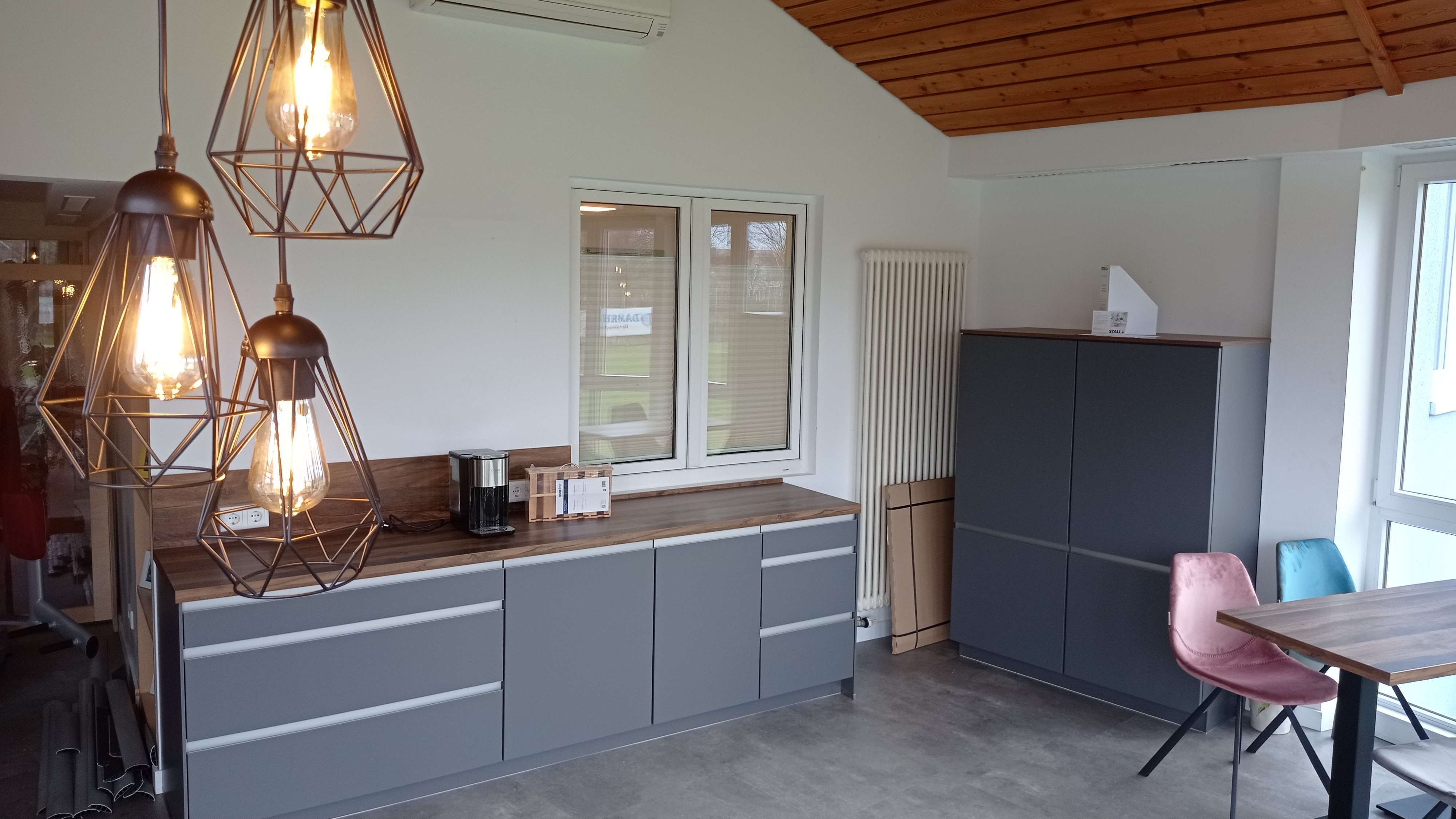 Moderne Küche mit hölzerner Arbeitsplatte, grauen Schränken, Pendelleuchten und einem Tisch mit bunten Stühlen.