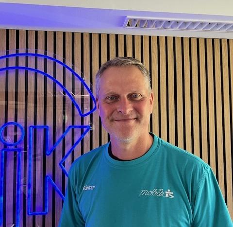 Mann mit kurzem Haar lächelt vor einer Holzvertäfelung und einem blauen Logo im Hintergrund. Er trägt ein türkisfarbenes Shirt.