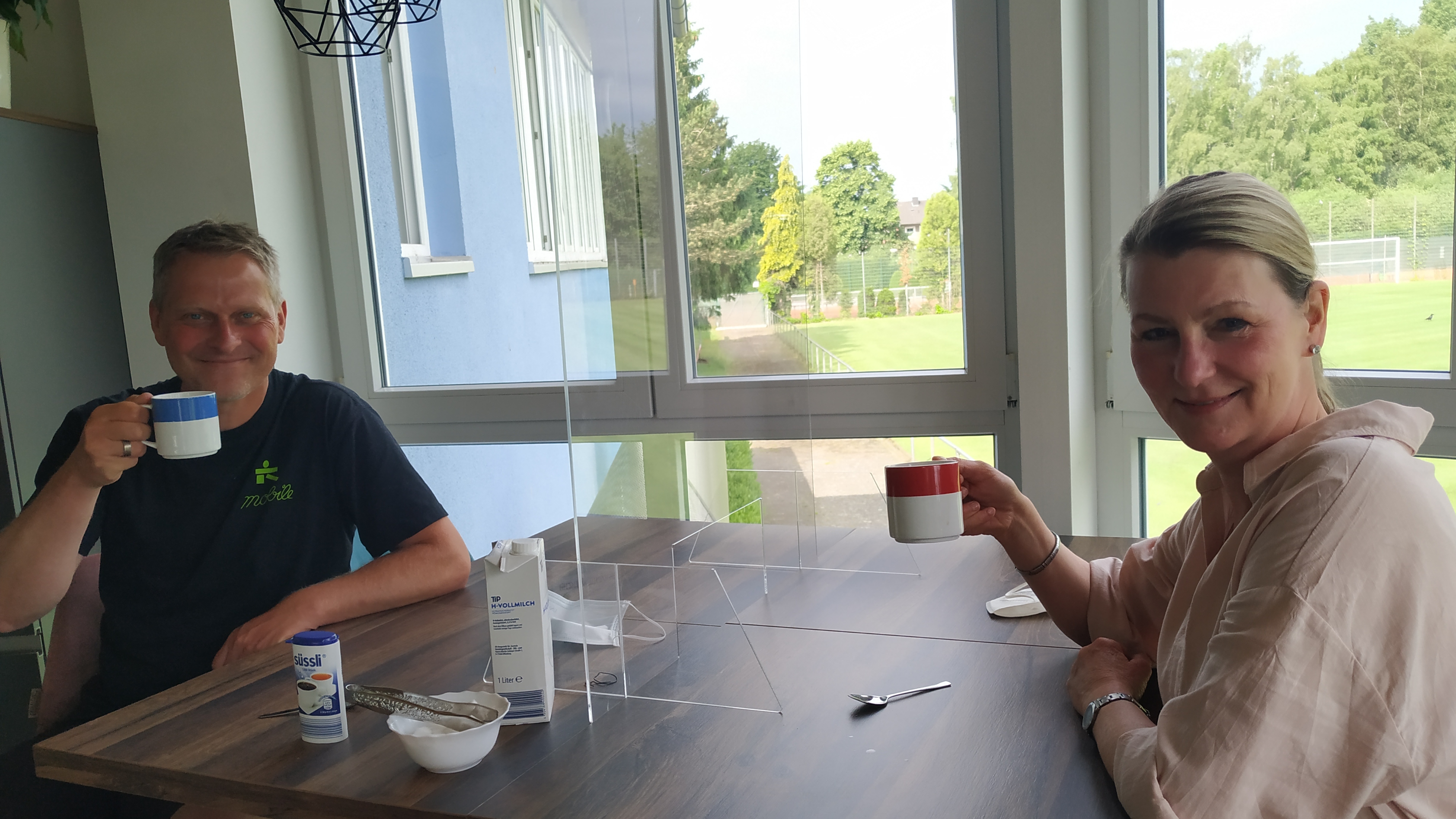 Zwei Personen sitzen an einem Tisch und halten Tassen; im Hintergrund sieht man ein Sportfeld durch ein Fenster.