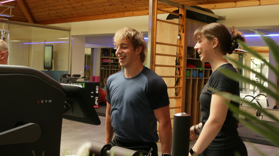 Zwei Personen lächeln und stehen vor einem Fitnessgerät in einem modernen Trainingsraum mit Holzdecke.