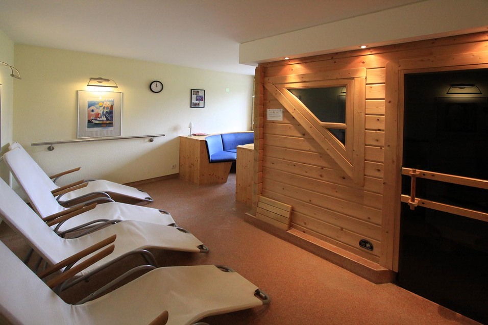Ruhebereich mit Liegen, Holzwand, Sauna, Tisch und Sitzbank in entspannter Atmosphäre. Wandbilder und Uhr vorhanden.