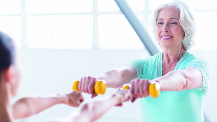 Ältere Frau im Fitnessstudio hebt gelbe Hanteln zusammen mit einer Trainerin in sportlicher Kleidung.
