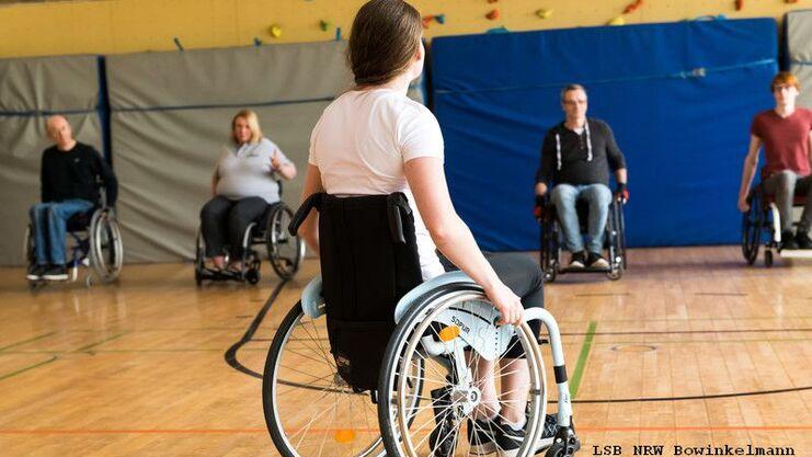 Fünf Personen im Rollstuhl sitzen in einer Sporthalle, eine Person ist von hinten sichtbar. Fokus auf die aktive Teilnahme.