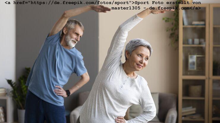 Zwei ältere Menschen machen im Wohnzimmer Dehnübungen, eine Frau beugt sich zur Seite, ein Mann steht im Hintergrund.