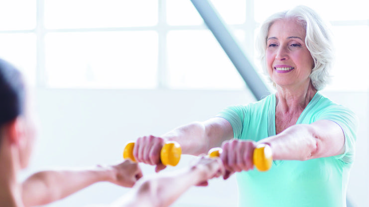 Ältere Frau im Fitnessstudio hebt gelbe Hanteln zusammen mit einer Trainerin in sportlicher Kleidung.