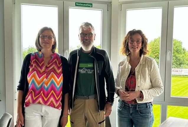 Drei Personen stehen vor großen Fenstern mit Blick auf ein grünes Feld, im Hintergrund ein Hinweisschild „Notausstieg“.