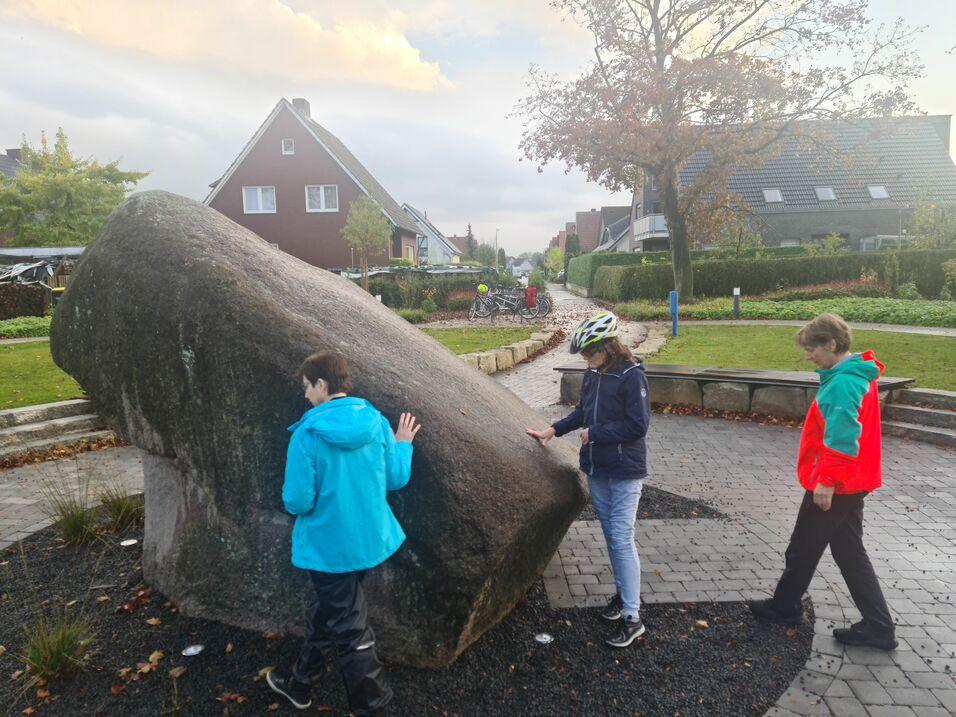 Drei Kinder erkunden einen großen Felsen in einem Park, umgeben von Gebäuden und Grünflächen.