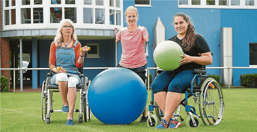 Drei Frauen in Rollstühlen posieren auf einer Wiese, umgeben von Fitnessbällen in verschiedenen Farben.