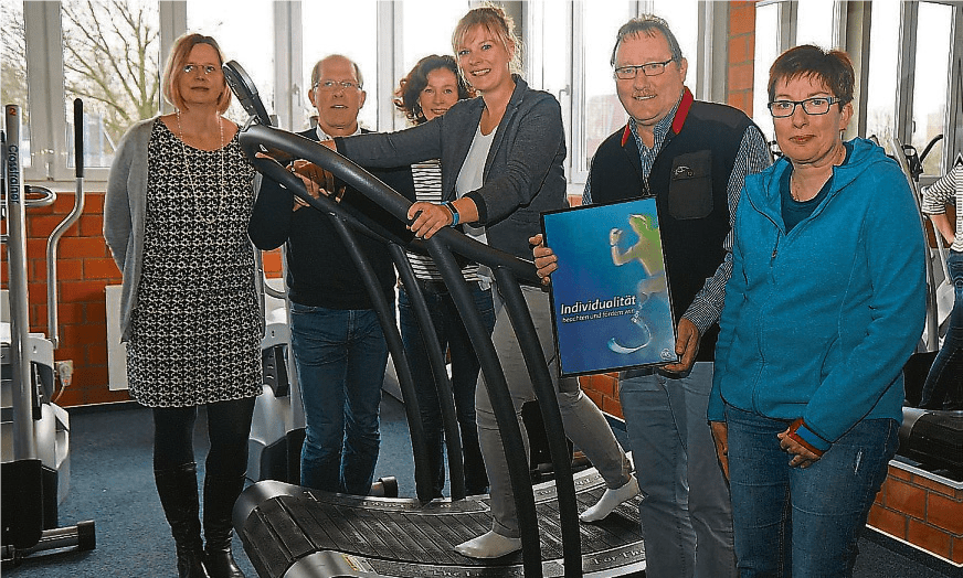 Gruppe von fünf Personen, die auf einem Fitnessgerät stehen, während eine Frau ein Plakat mit dem Titel „Individualität“ hält.