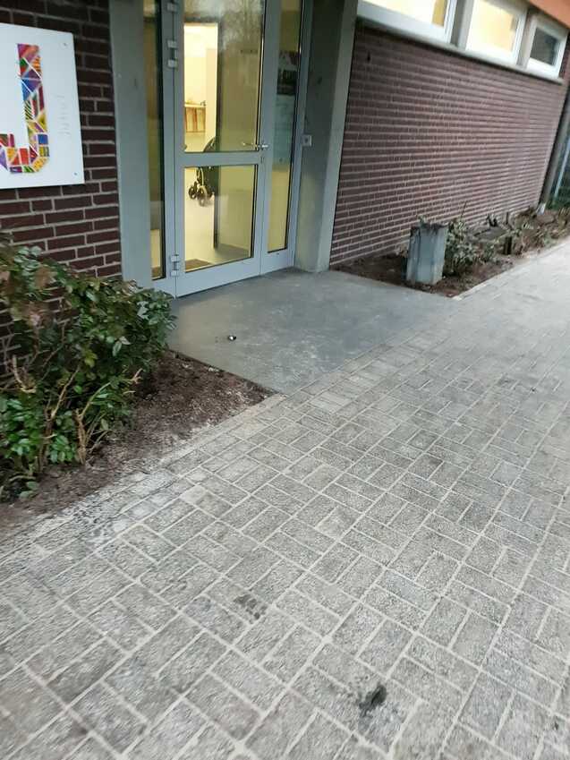 Eingang einer Einrichtung mit Glastür, umgeben von Pflanzen und einem gepflasterten Weg. Innenraum unscharf im Hintergrund sichtbar.
