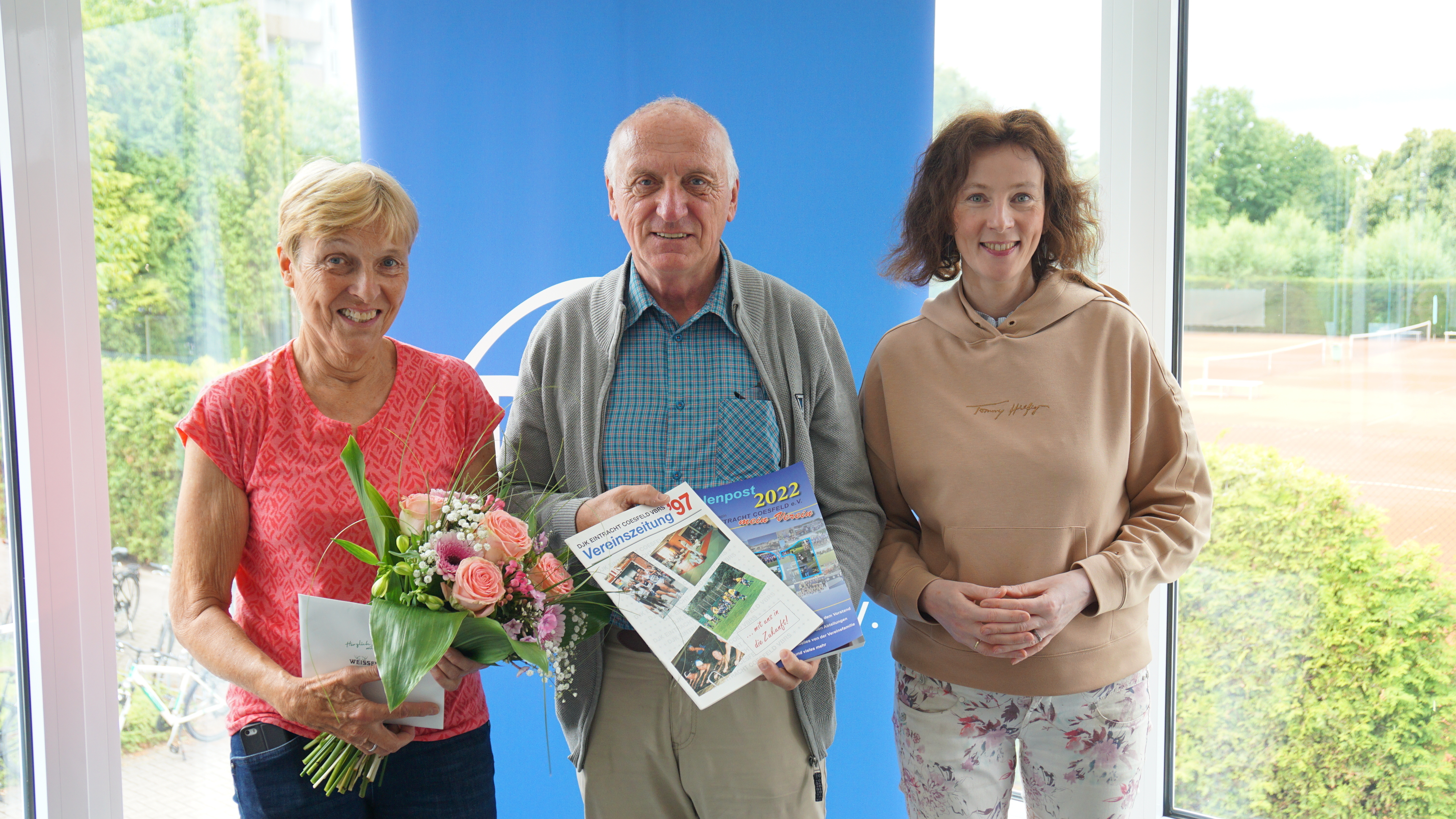 Drei Personen stehen vor einem blauen Hintergrund und halten Blumen sowie zwei Vereinszeitungen in den Händen.