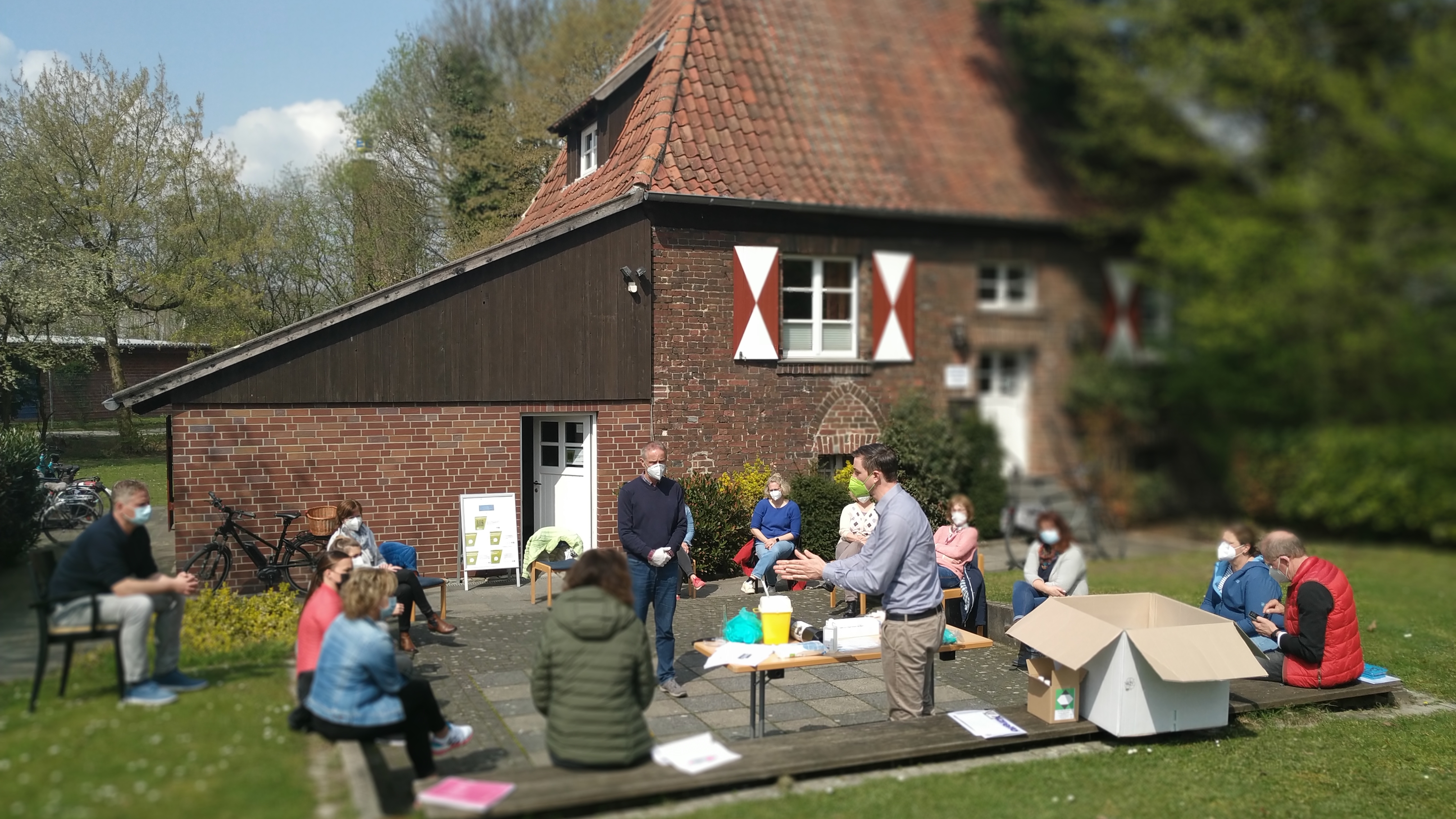 Gruppe von Menschen sitzt vor einem Fachwerkhaus und hört einem Dozenten im Freien aufmerksam zu.