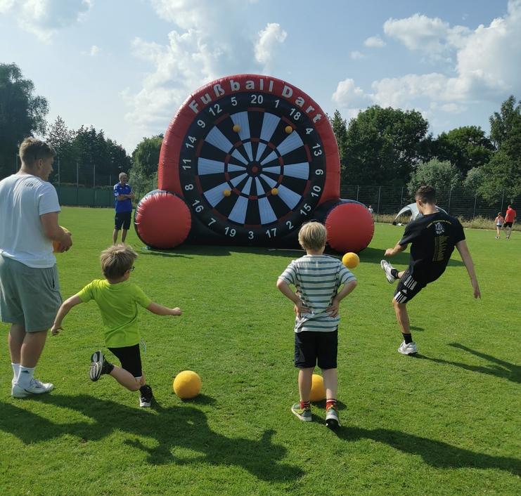 Vier Kinder und ein Erwachsener spielen Fußball-Dart mit einem großen, aufblasbaren Dartboard auf einer grünen Wiese.