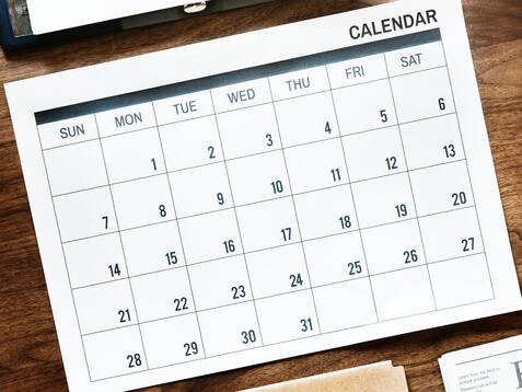 Blick auf einen Tisch mit einem Kalender, Laptop, Smartphone, Notizblock, Uhr und einer Zeitung.