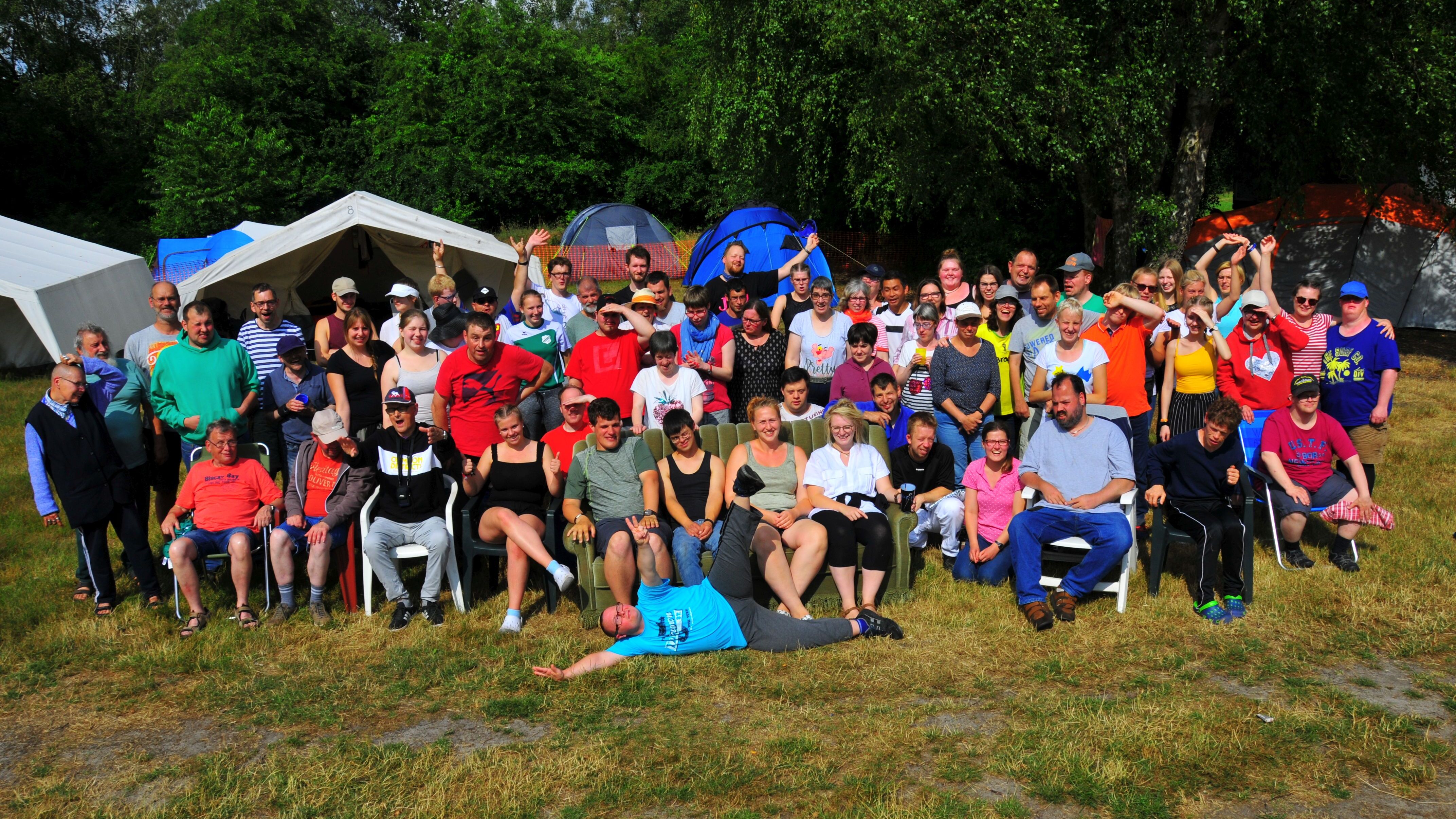 Gruppenfoto von 65 Personen im Freien, umgeben von Zelten und Bäumen, einige winken und lachen.