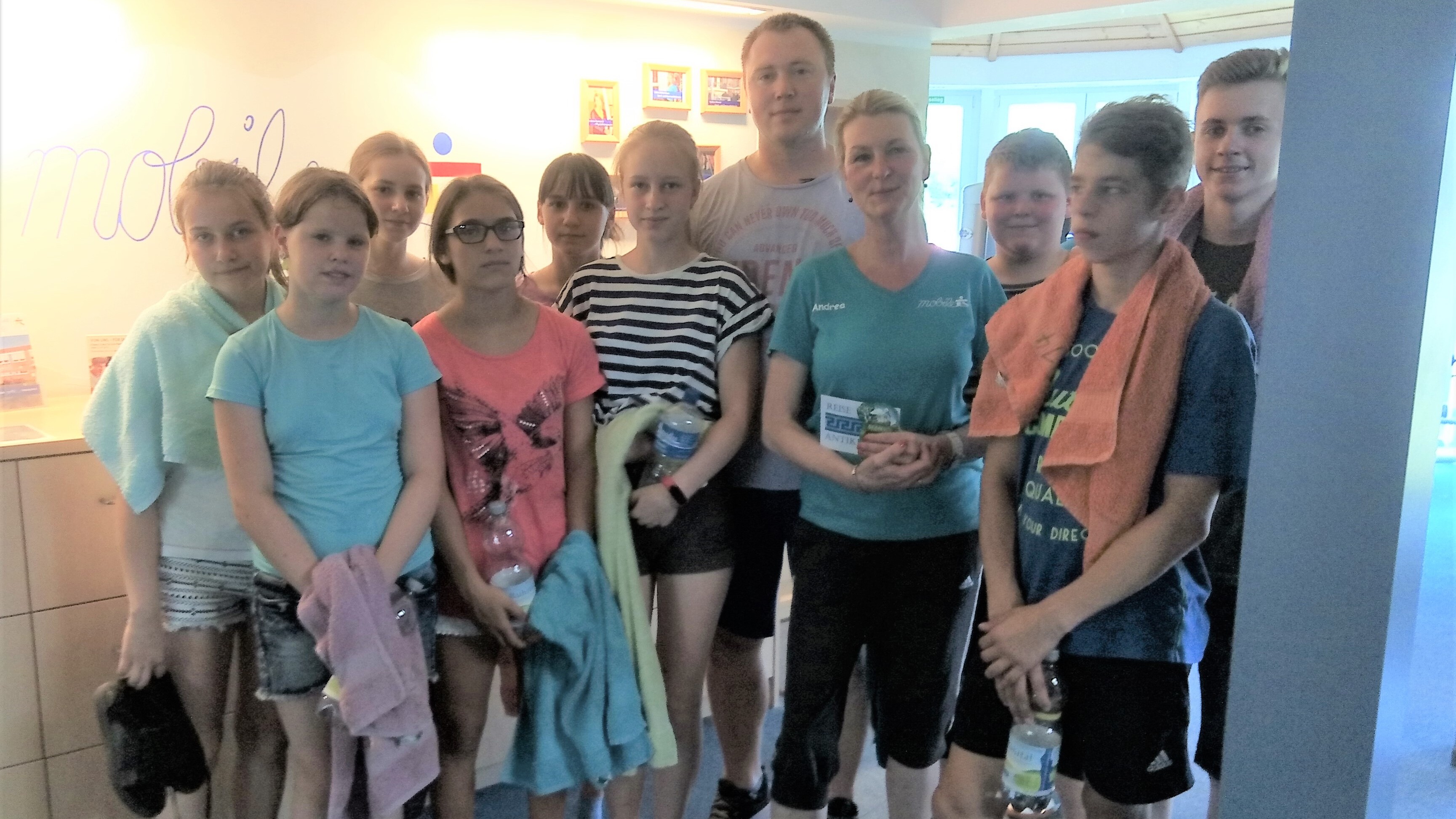 Gruppe von zwölf Jugendlichen und einem Erwachsenen in Sportkleidung, lächelnd vor einer Wand mit Informationsmaterial.